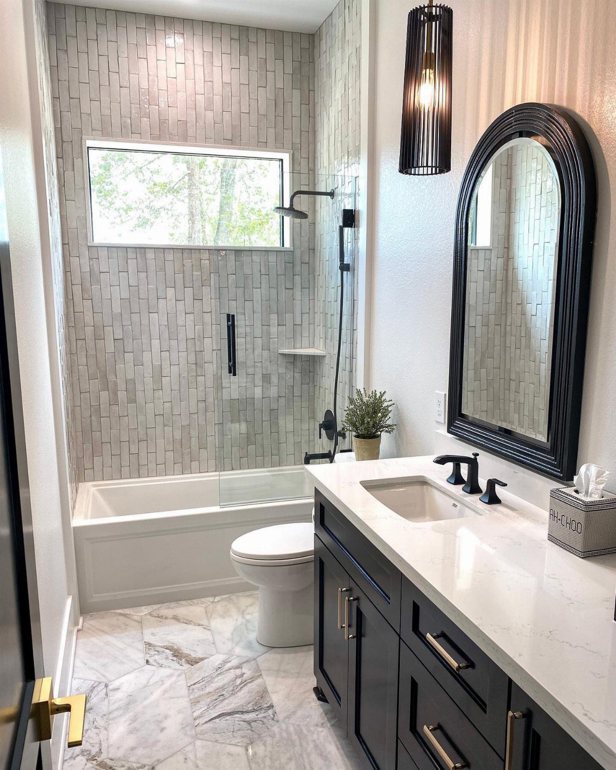 A narrow bathroom with a white bathtub, shower, toilet, and dark vanity with an arched mirror.