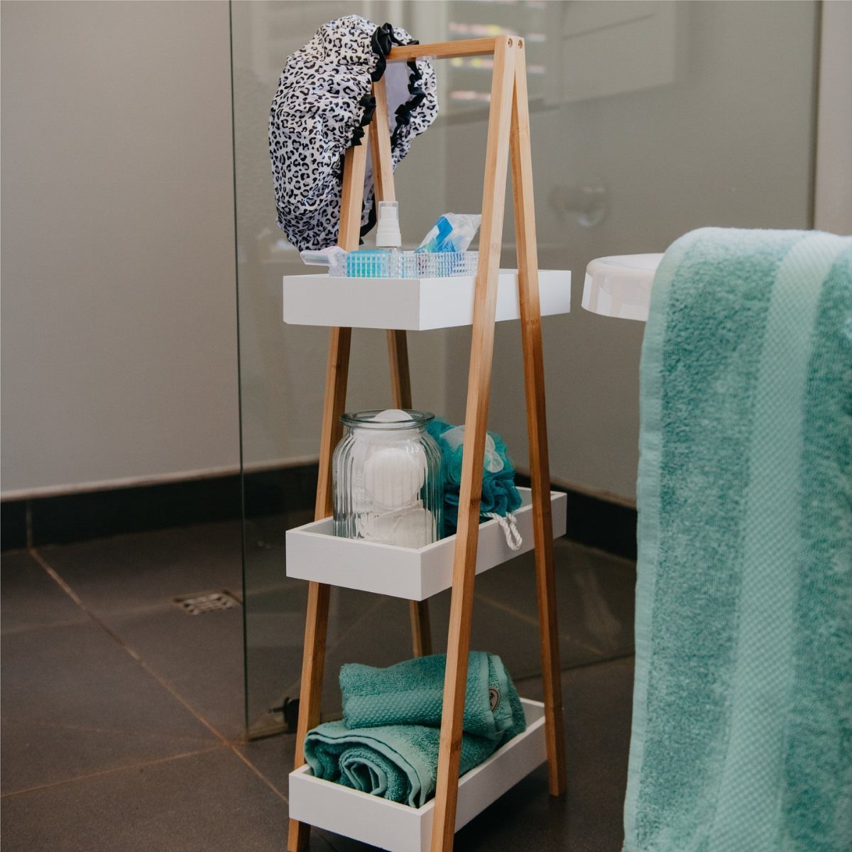A wooden ladder shelf holding folded teal towels and bathroom toiletries in a modern bathroom.