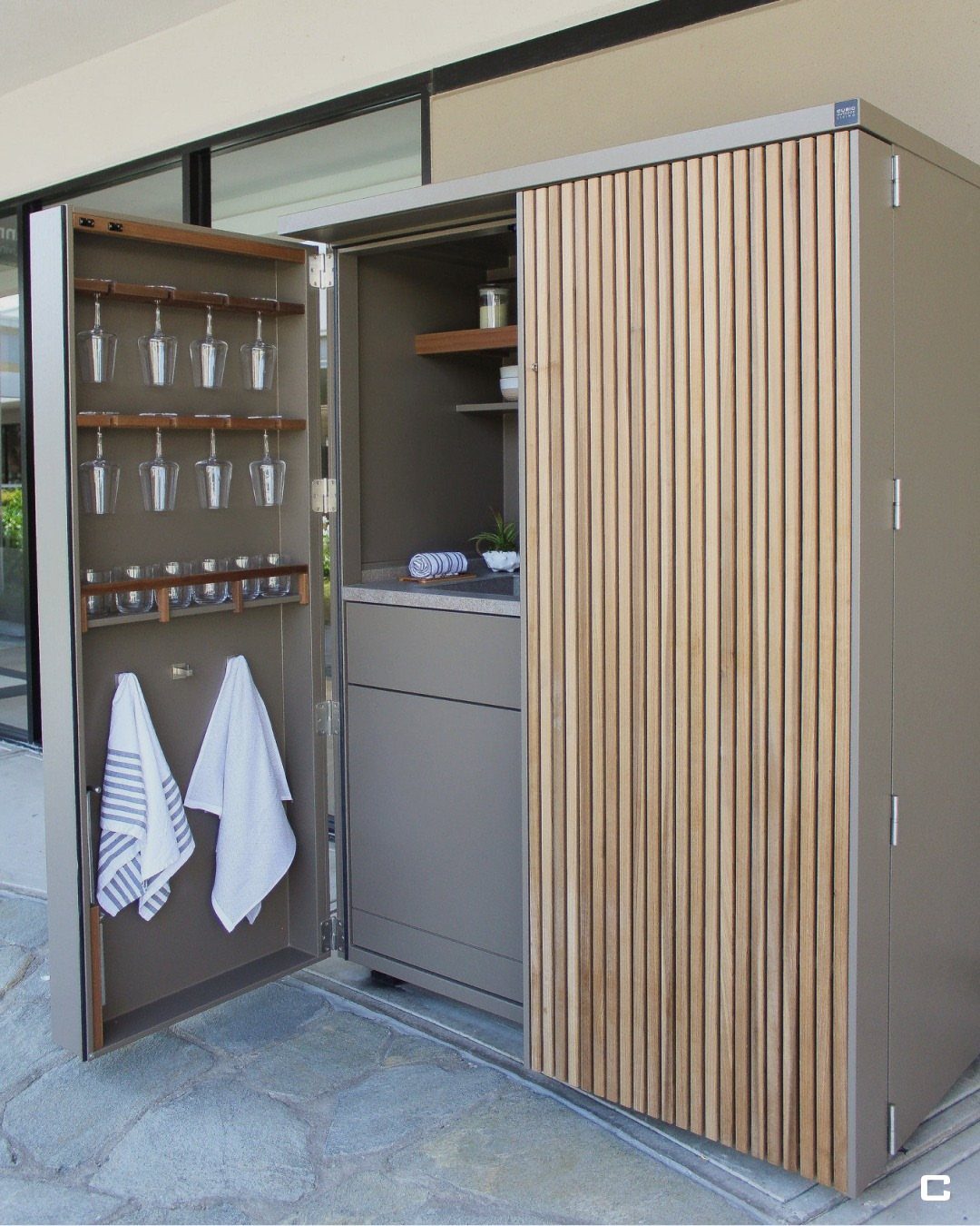 An open outdoor storage cabinet containing glassware, hanging stemware, and white towels on a stone patio.