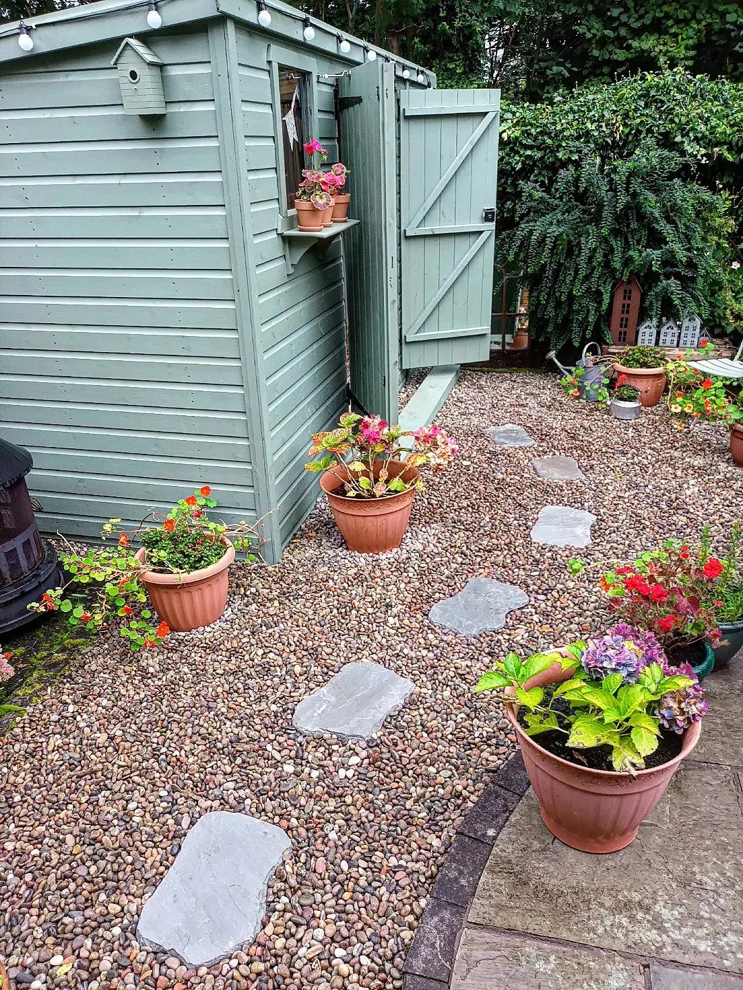 A small green wooden garden shed with an open door surrounded by potted flowers and gravel.