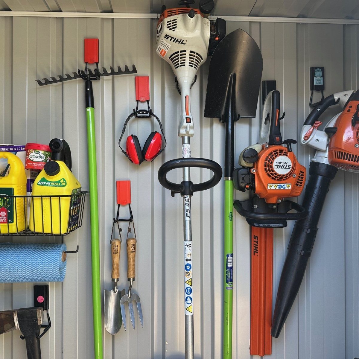 Garden tools like a rake, shovel, and leaf blower hanging organized on a grey shed wall.