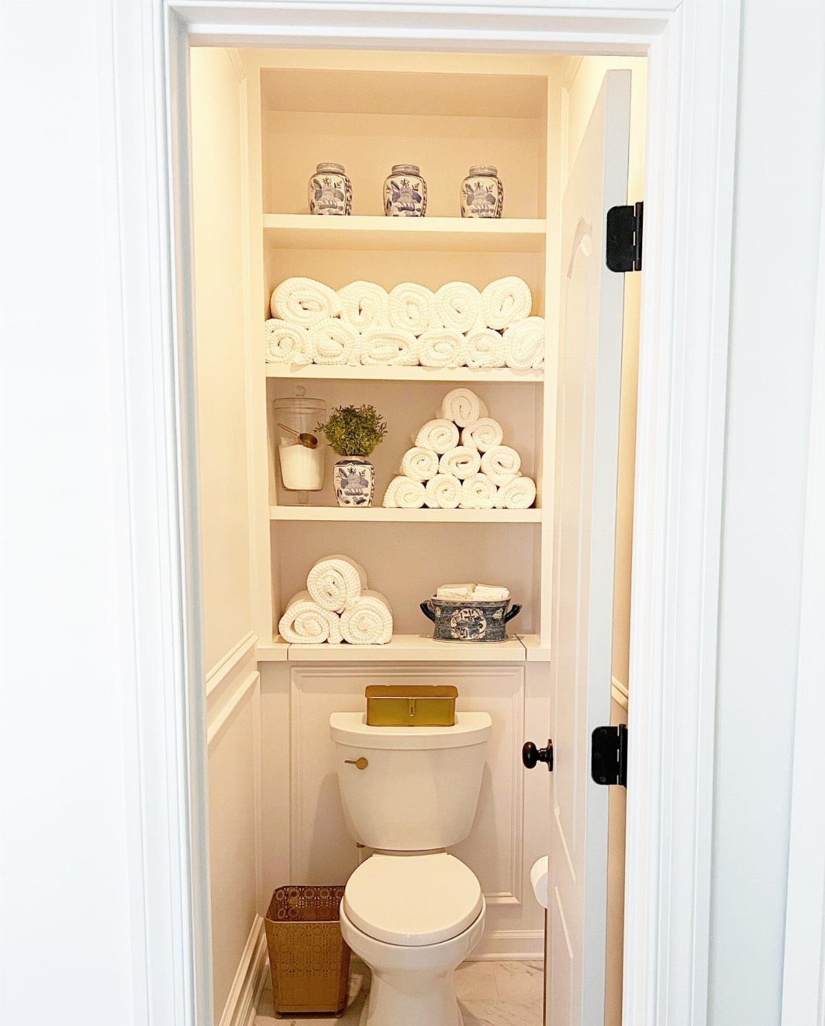 Organized white shelves filled with rolled towels and decorative jars above a toilet in a small bathroom.